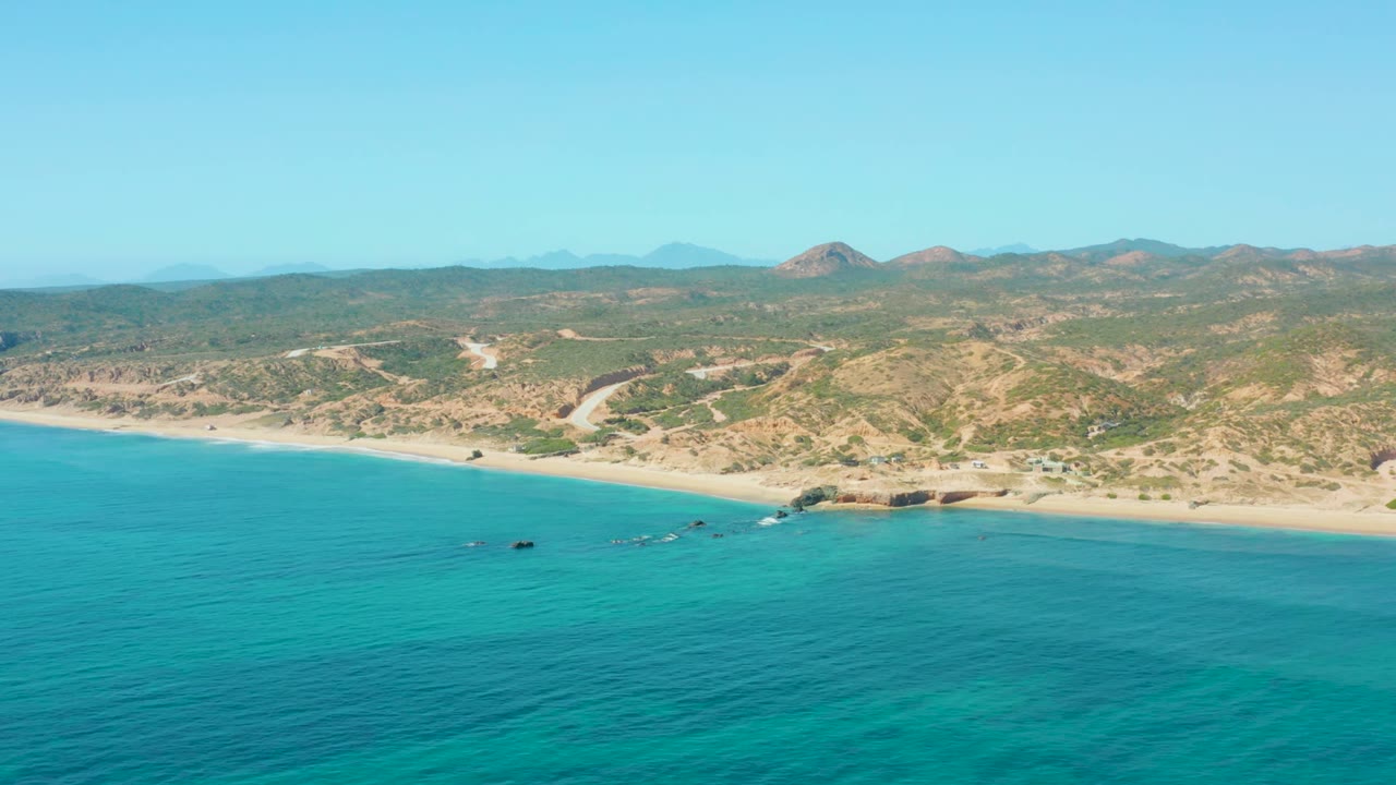 Aerial frame over El Tule Los Cabos with turquoise water in the foreground, a sandy beach across the middle, low dry hills beyond, and no visible people.