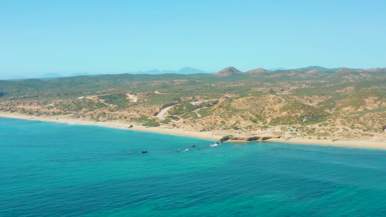 Aerial view at El Tule Los Cabos showing calm turquoise ocean below, a pale beach running left to right, desert hills in back, and no visible people.
