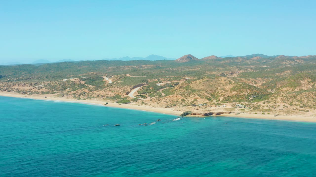 The El Tule Los Cabos shoreline appears with a wide band of turquoise sea, a light sand beach, scrub-covered hills, faint dirt tracks, and no visible people.