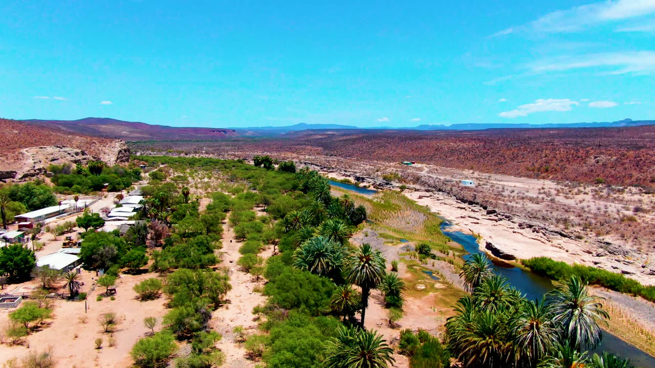 Aerial frame over San Juanico, Baja California Sur showing a narrow river, palm clusters, sandy ground, reddish hills, distant mountains, blue sky, and no visible people.
