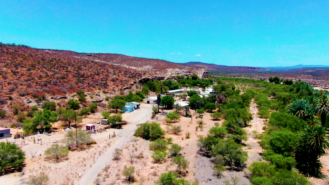 Aerial view over San Juanico, Baja California Sur, with sandy terrain, scattered green trees, palms, small buildings, low hills, and no visible people.