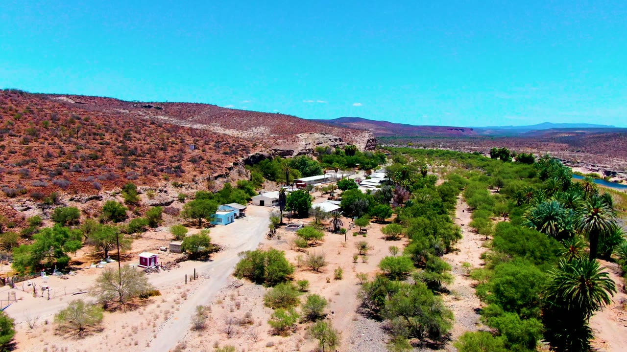 Drone view of sandy desert, palms, scattered buildings, and a road near low hills.