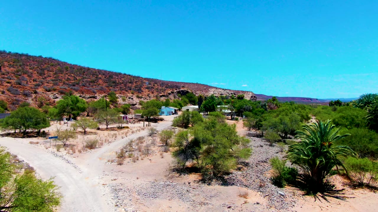 Opening frame in San Juanico, Baja California Sur, with a pale dirt road, sparse desert trees, small structures, a palm, a reddish hillside, and no visible people.