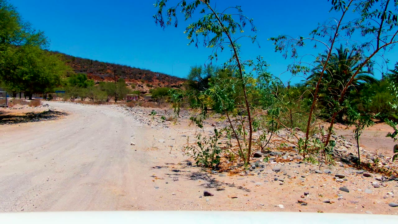 Start of the San Juanico road clip, showing a pale dirt track, rocky ground, foreground scrub, low hills, clear blue sky, and no visible people.