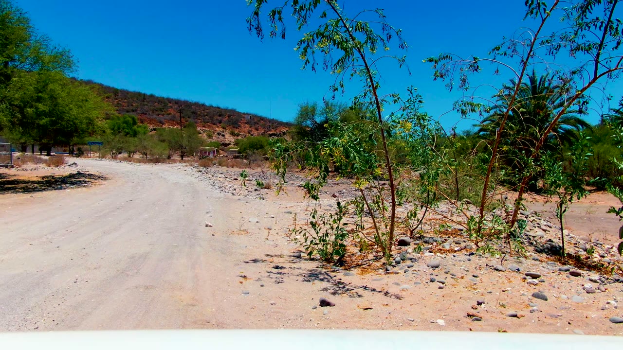 At 0:02, the San Juanico dirt road passes rocky shoulders and sparse trees, with small distant structures partly visible near the left side and no visible people.