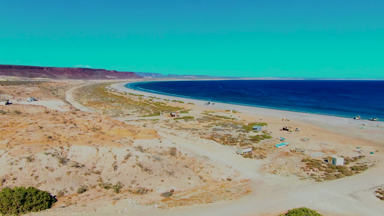 Opening aerial frame over San Juanico, Baja California Sur, showing sandy dunes, sparse scrub, a pale beach, dark blue ocean, and no visible people.