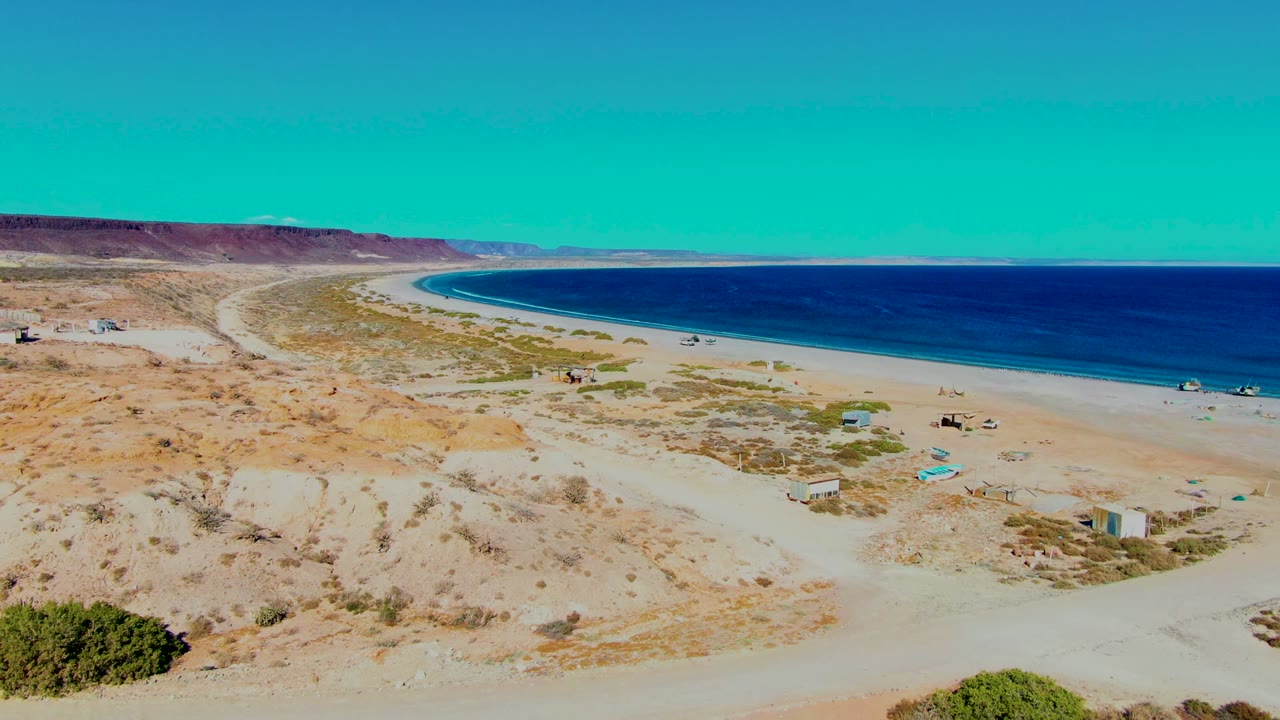 Aerial frame in San Juanico with a sandy track crossing the foreground, scrub patches on the dunes, small beach structures, a curved shoreline, and no visible people.