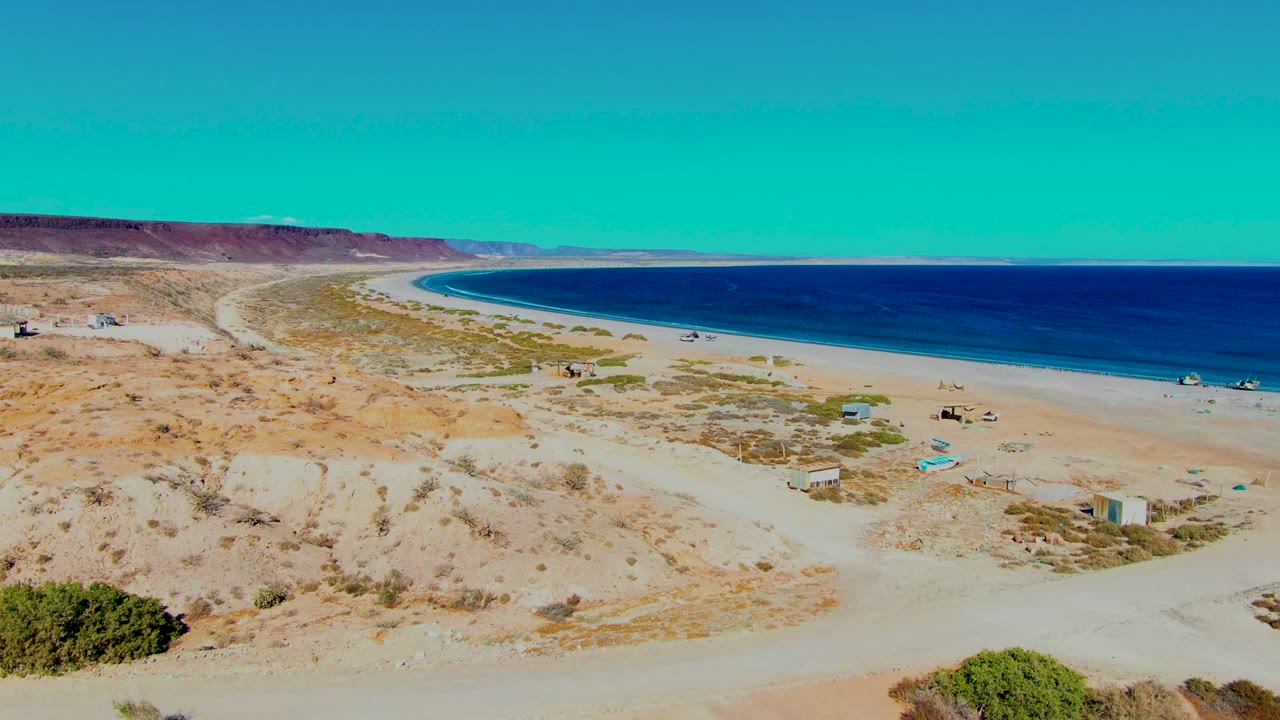 The frame looks across San Juanico's desert shoreline, with pale sand, scattered vegetation, small rectangular structures near the beach, deep blue water, and no visible people.
