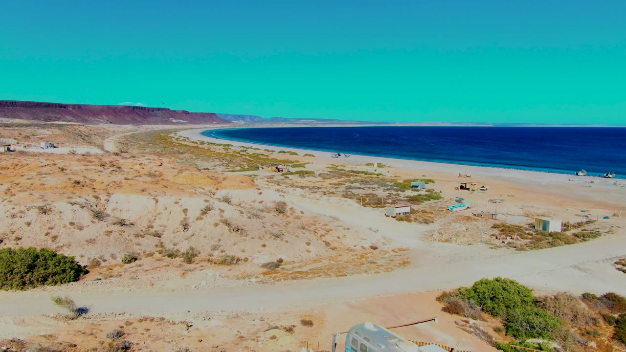 Wide elevated view of San Juanico coastal desert with pale dunes, sparse shrubs, blue ocean, distant mountains, and no visible people.