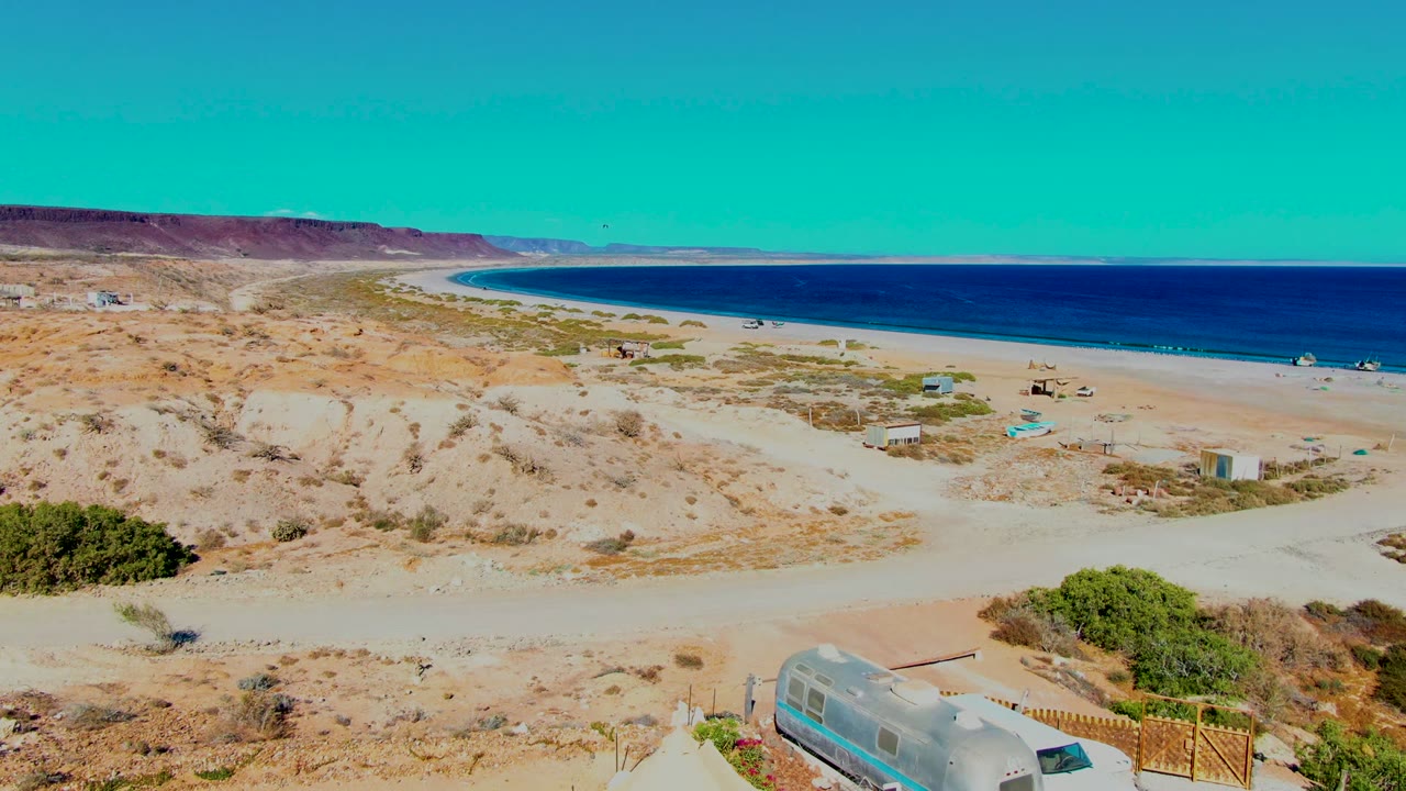 San Juanico beach desert frame with ocean across the upper right, tan dunes through the center, small structures near the sand, and no visible people.