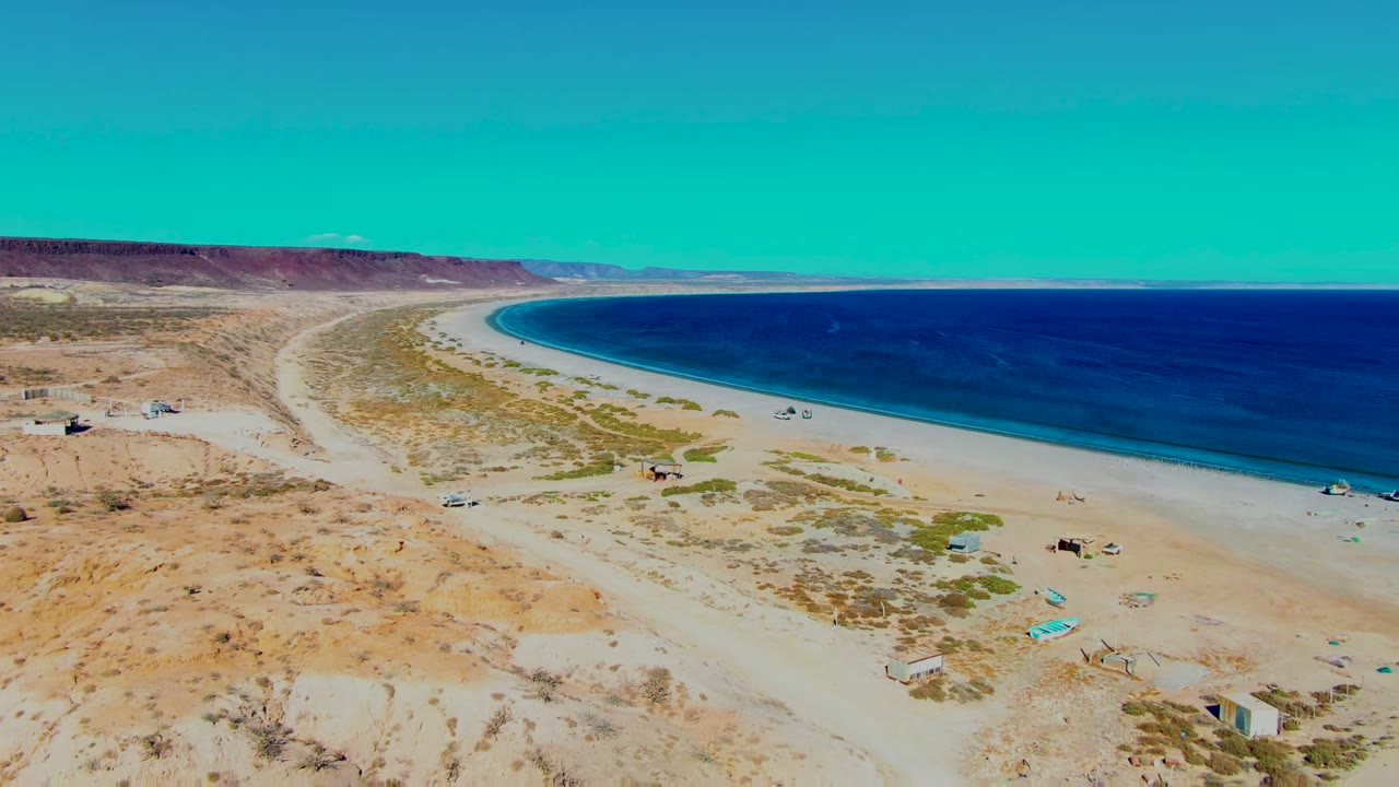 Aerial frame over San Juanico, Baja California Sur, Mexico, showing pale desert sand, a curving beach, deep blue ocean, scattered vehicles, and no visible people.