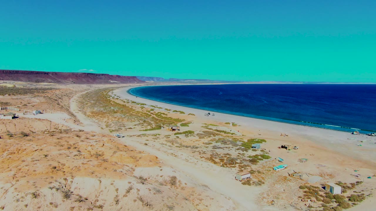 The aerial view looks along the San Juanico beach with dark blue water on the right, arid sand in the foreground, vehicles near the beach, and no visible people.