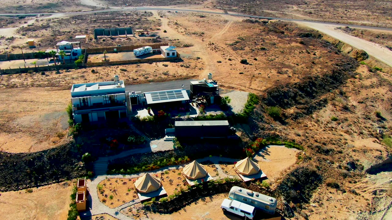 Wide aerial view in San Juanico, Baja California Sur showing a desert property with solar-roofed buildings, canvas tents, a road above the site, a white van near the lower driveway, and no visible people.