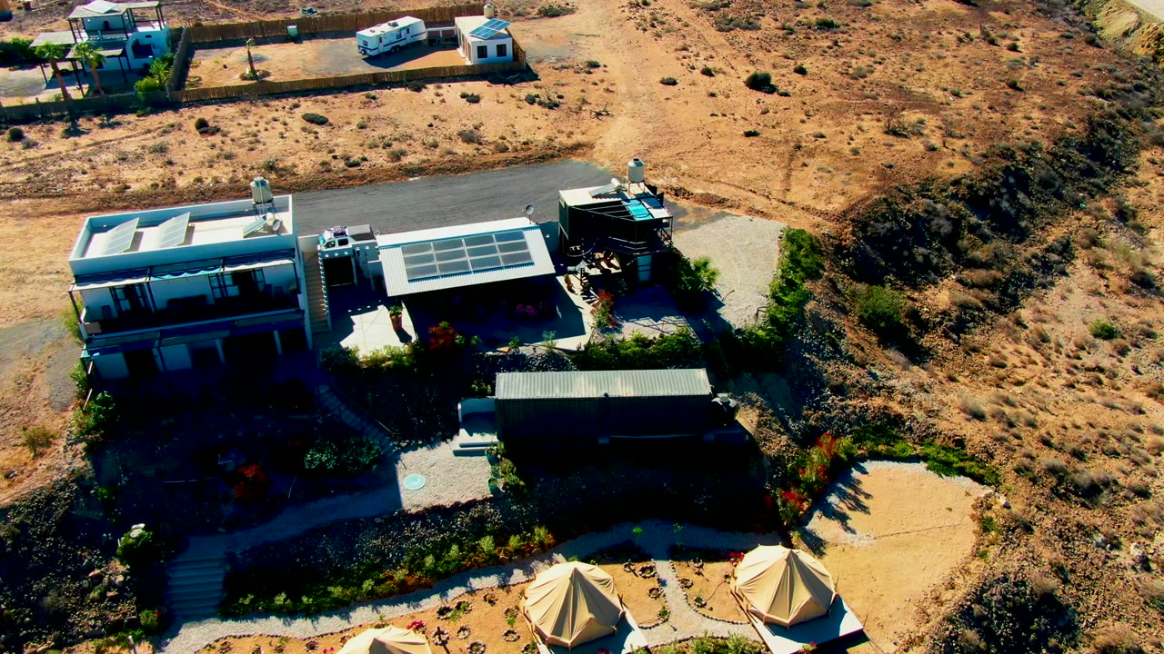 Overhead frame in San Juanico, Baja California Sur showing the van near the upper paved area, a solar-panel roof centered below it, canvas tents at the lower edge, surrounding sandy slopes, and no visible people.