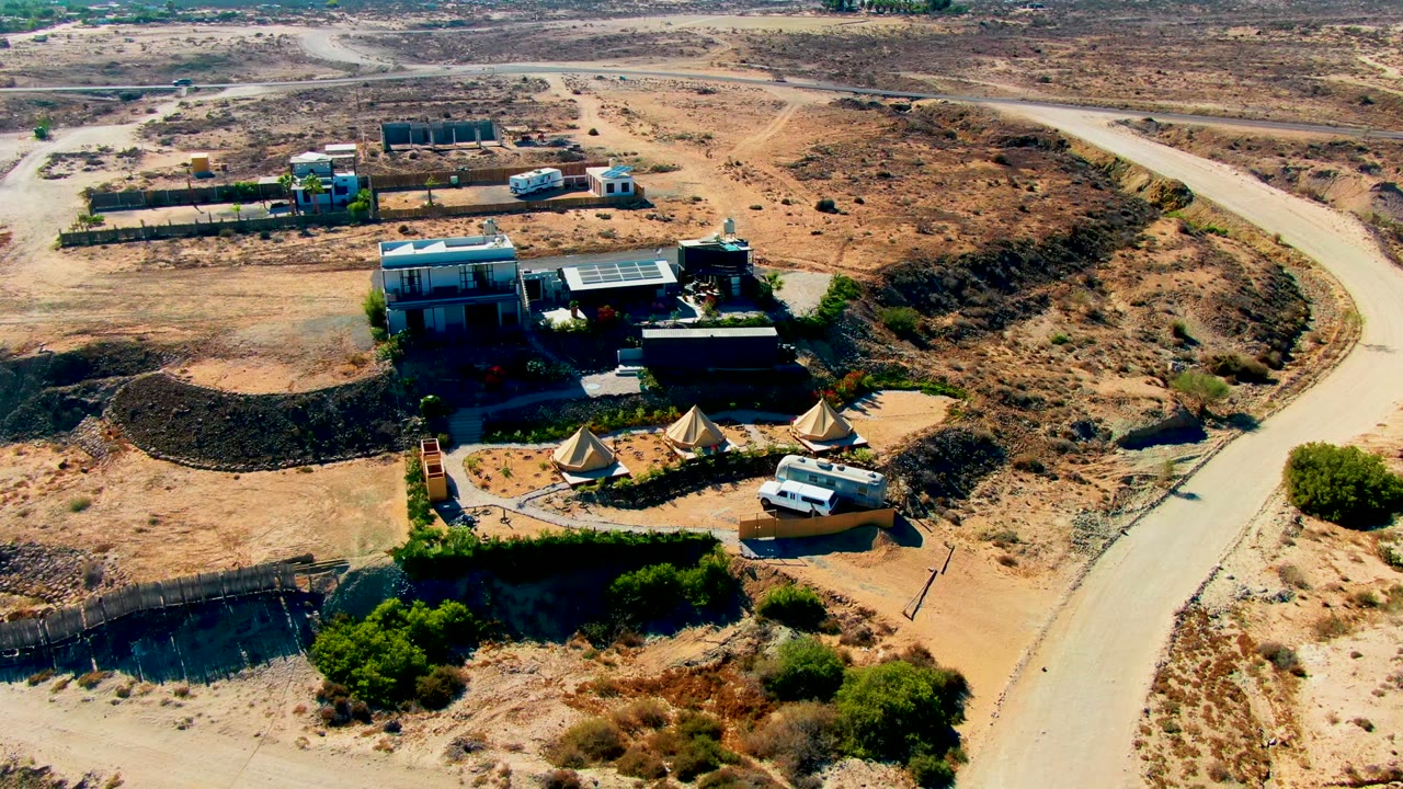Aerial frame over San Juanico, Baja California Sur showing low buildings, conical tents, a white van, dry scrubland, and a curved road at the start of the clip with no visible people.