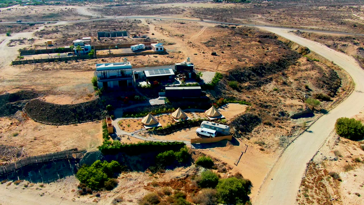 Drone-style overhead frame of the San Juanico desert property with buildings and tents in the lower center, the road bending along the right edge, scattered structures beyond, and no visible people.