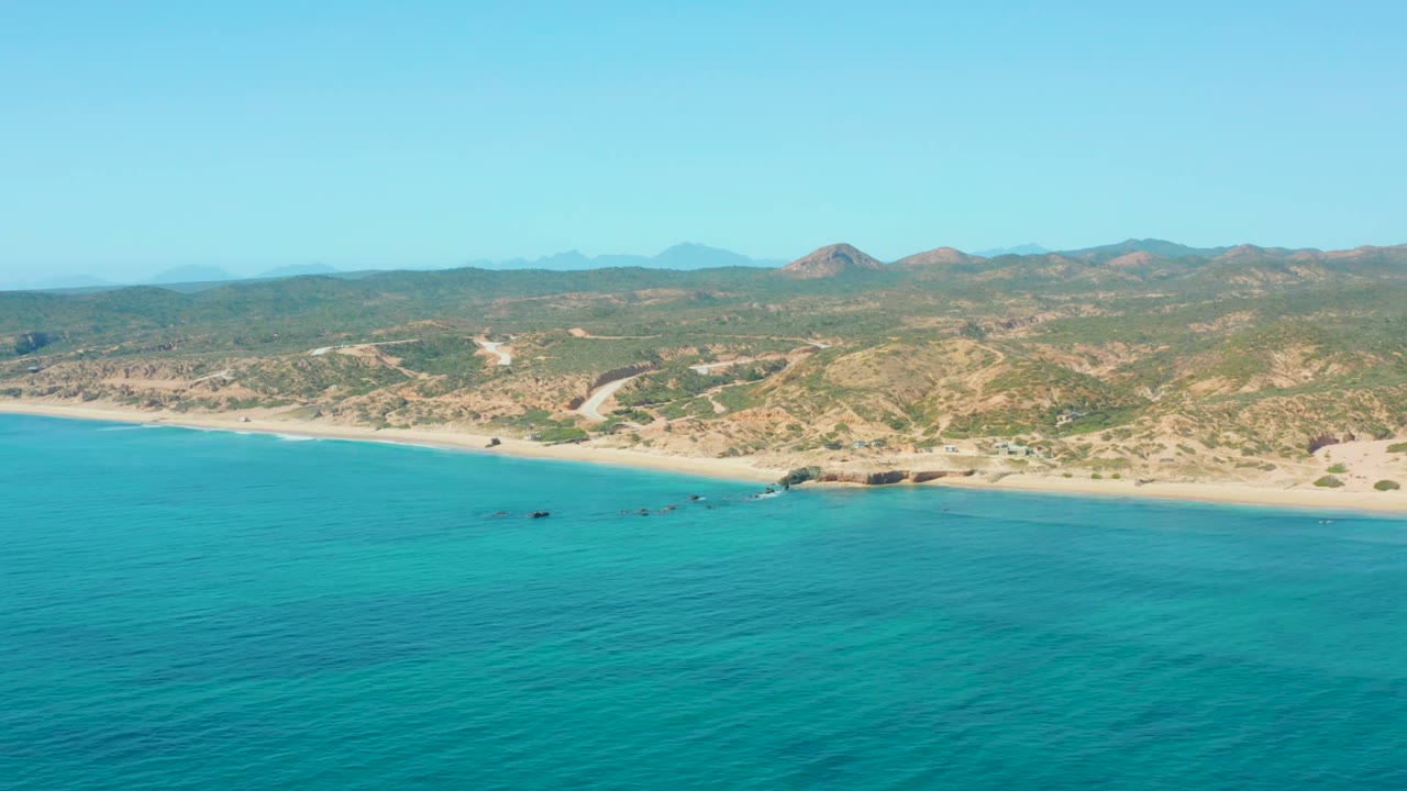 Opening aerial frame over El Tule Los Cabos with turquoise water in the foreground, a sandy shore beyond, small boats near the shallows, dry hills in back, and no visible people.