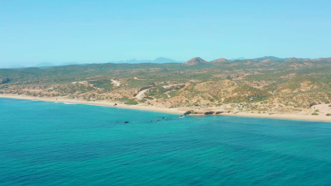 Wide aerial coastline with clear blue water, sand, boats, hills, and distant mountains.