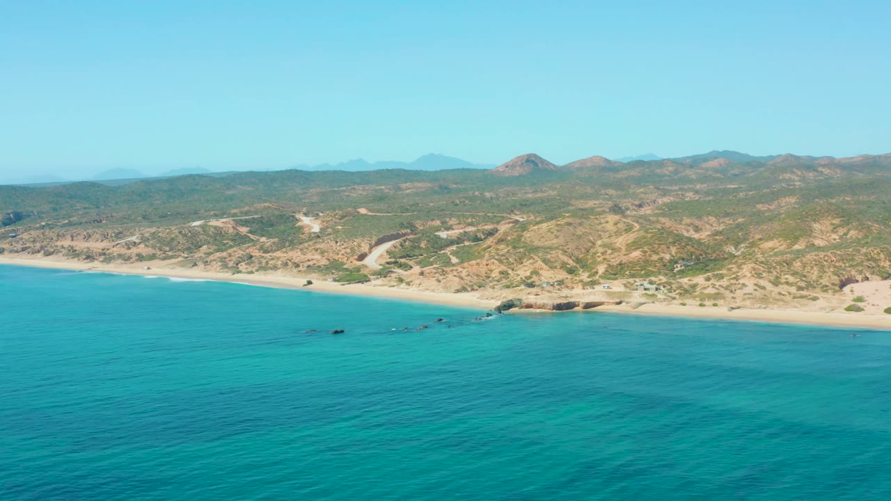 At 0:02, the aerial view shows a broad turquoise sea foreground, pale beach, scattered small boats near the coast, El Tule Los Cabos hills, and no visible people.
