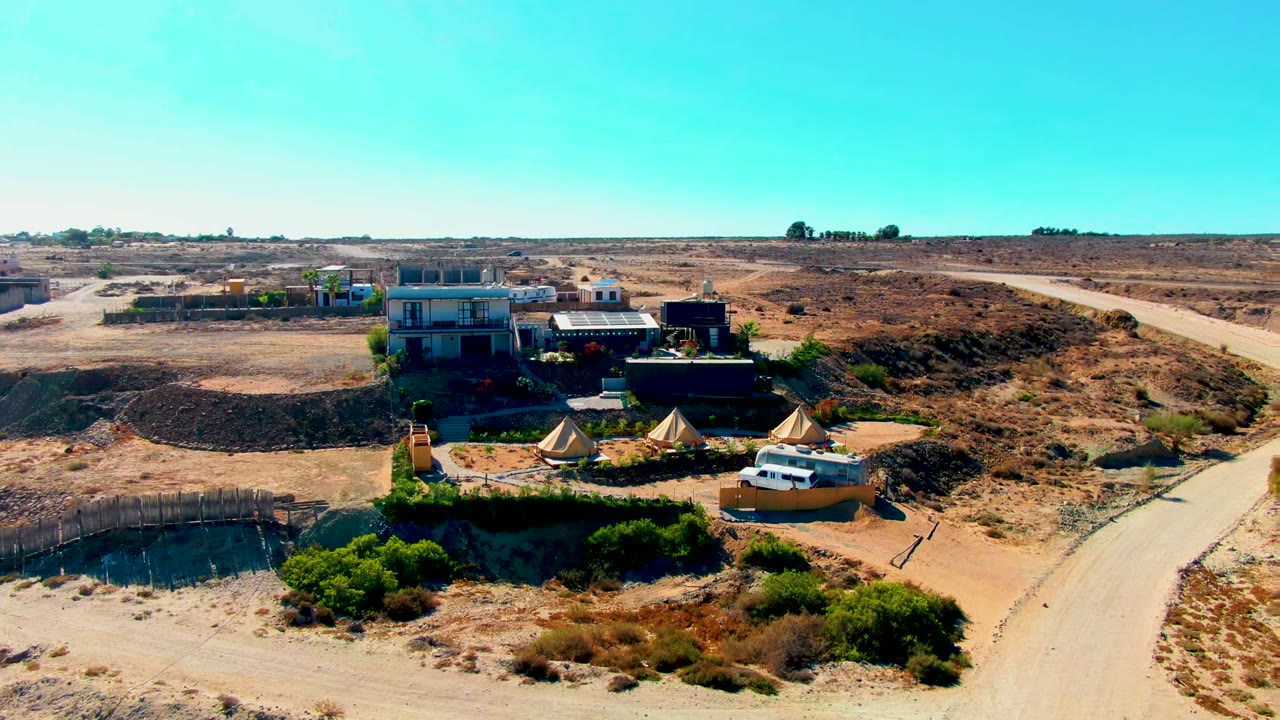 Wide aerial view over a San Juanico desert campsite with low buildings, canvas tents, a white van near the foreground road, dry scrubland, and no visible people.