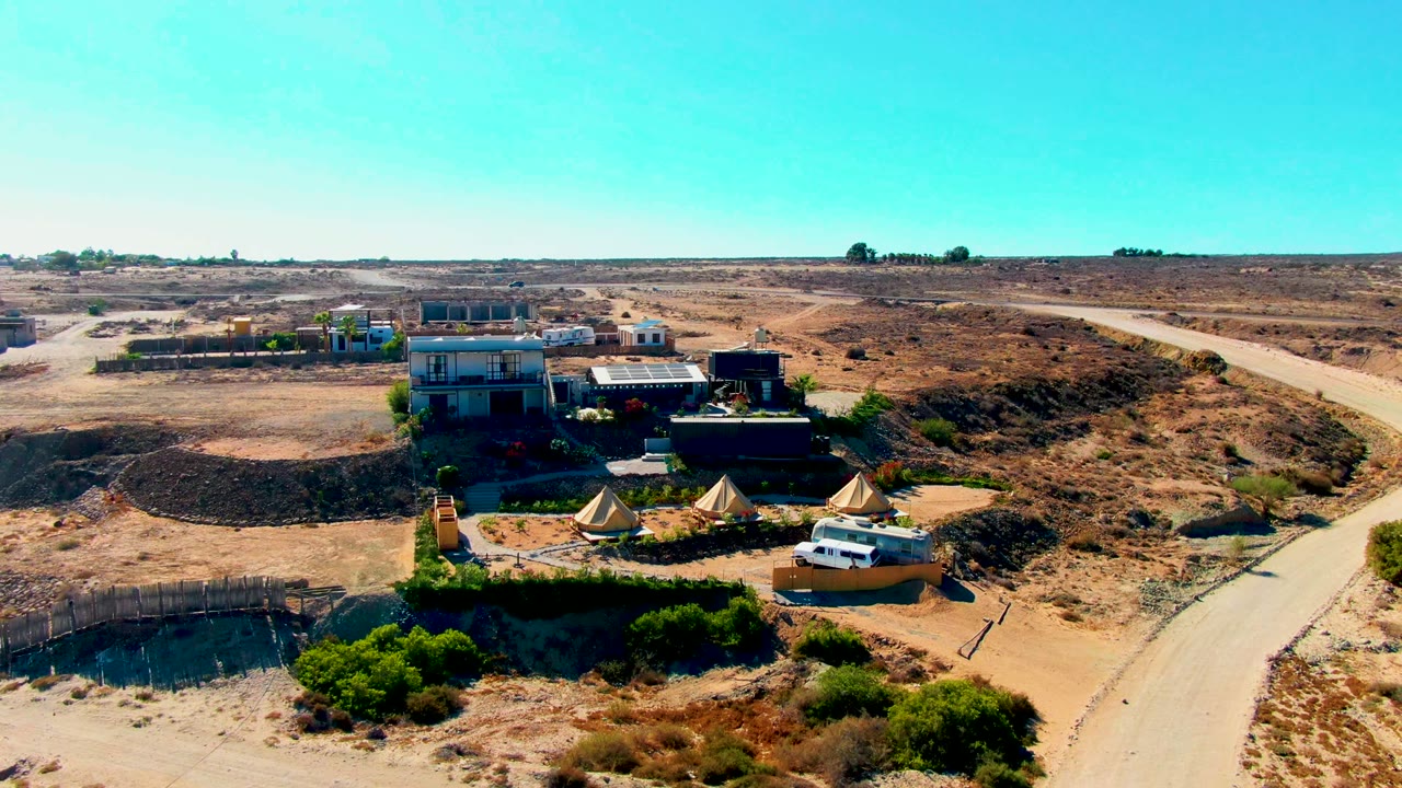 Drone view of white van beside tents and buildings in dry desert terrain.