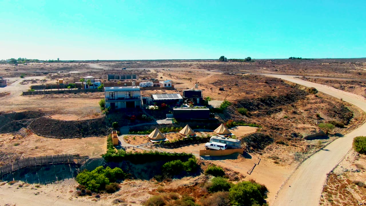 San Juanico aerial frame with a campsite cluster, white van, tan canvas tents, low buildings, dry brush, and a pale road crossing the right side, with no visible people.
