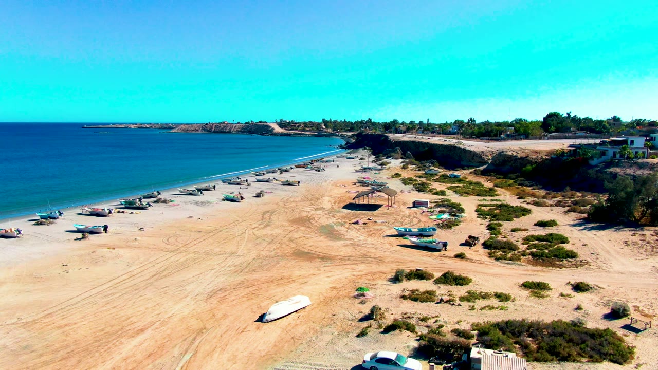 Drone-style view over San Juanico, Baja California Sur, with open sand in front, boats grouped near the waterline, calm ocean to the left, low coastal rocks beyond, and no visible people.