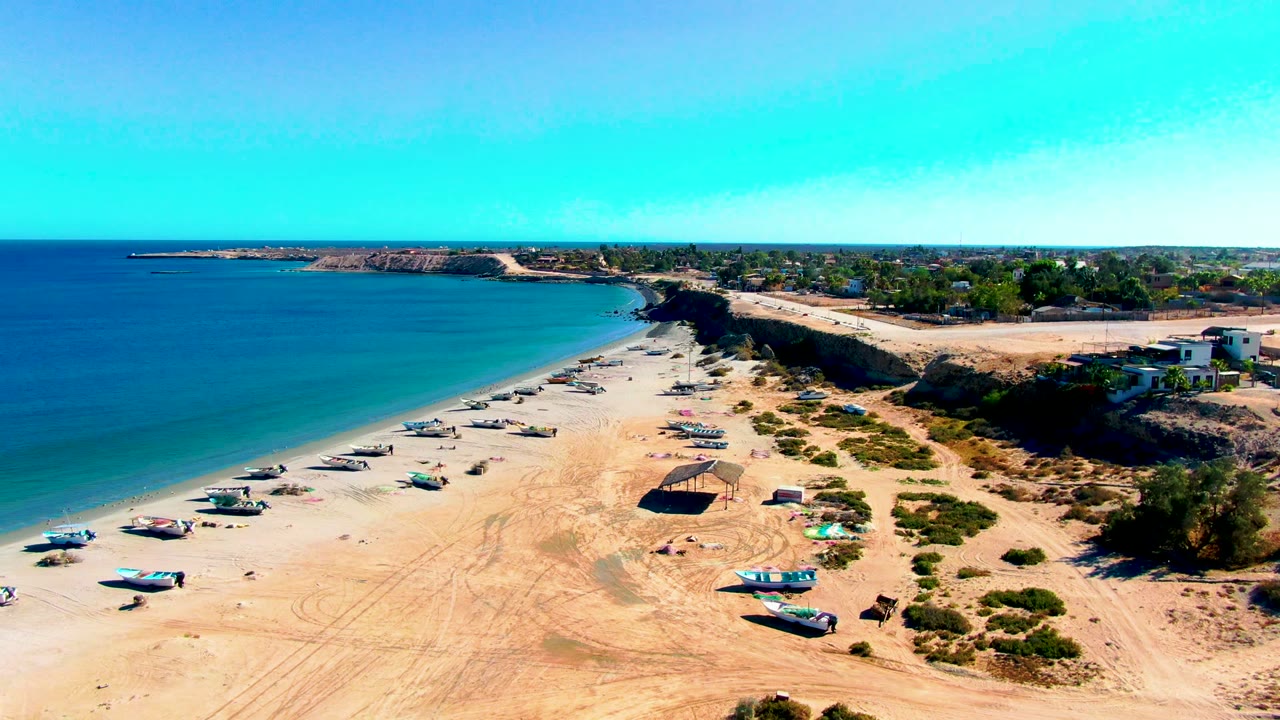 Wide aerial view of San Juanico beach with small boats lined along pale sand, blue ocean on the left, low cliffs and buildings on the right, and no visible people.
