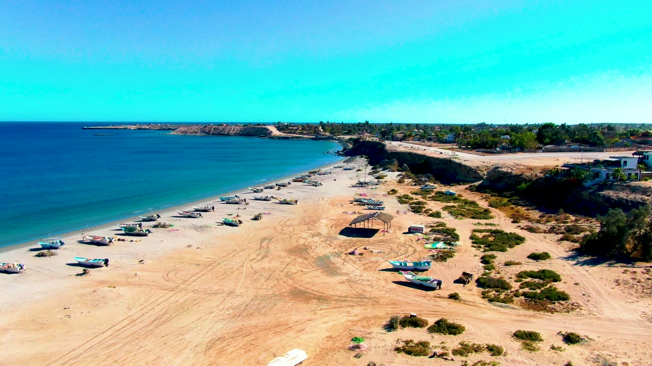 Elevated frame of San Juanico beach with calm blue water, rows of small boats on sand, scrub-covered dunes, low buildings near the bluff, and no visible people.