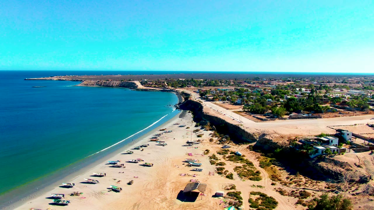 High aerial view over San Juanico, Baja California Sur, with blue ocean on the left, small boats on pale sand, low cliffs, inland buildings, and no visible people.