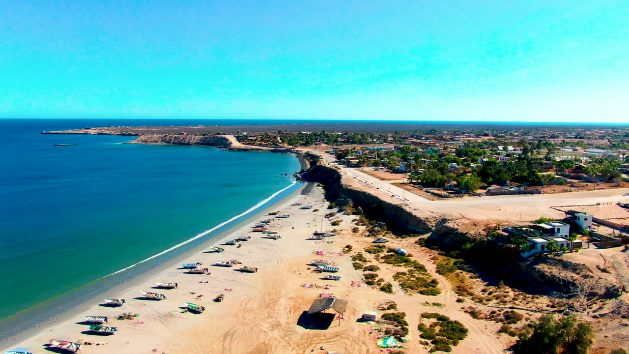 Drone view along a sandy shoreline with boats, turquoise water, cliffs, and houses inland.