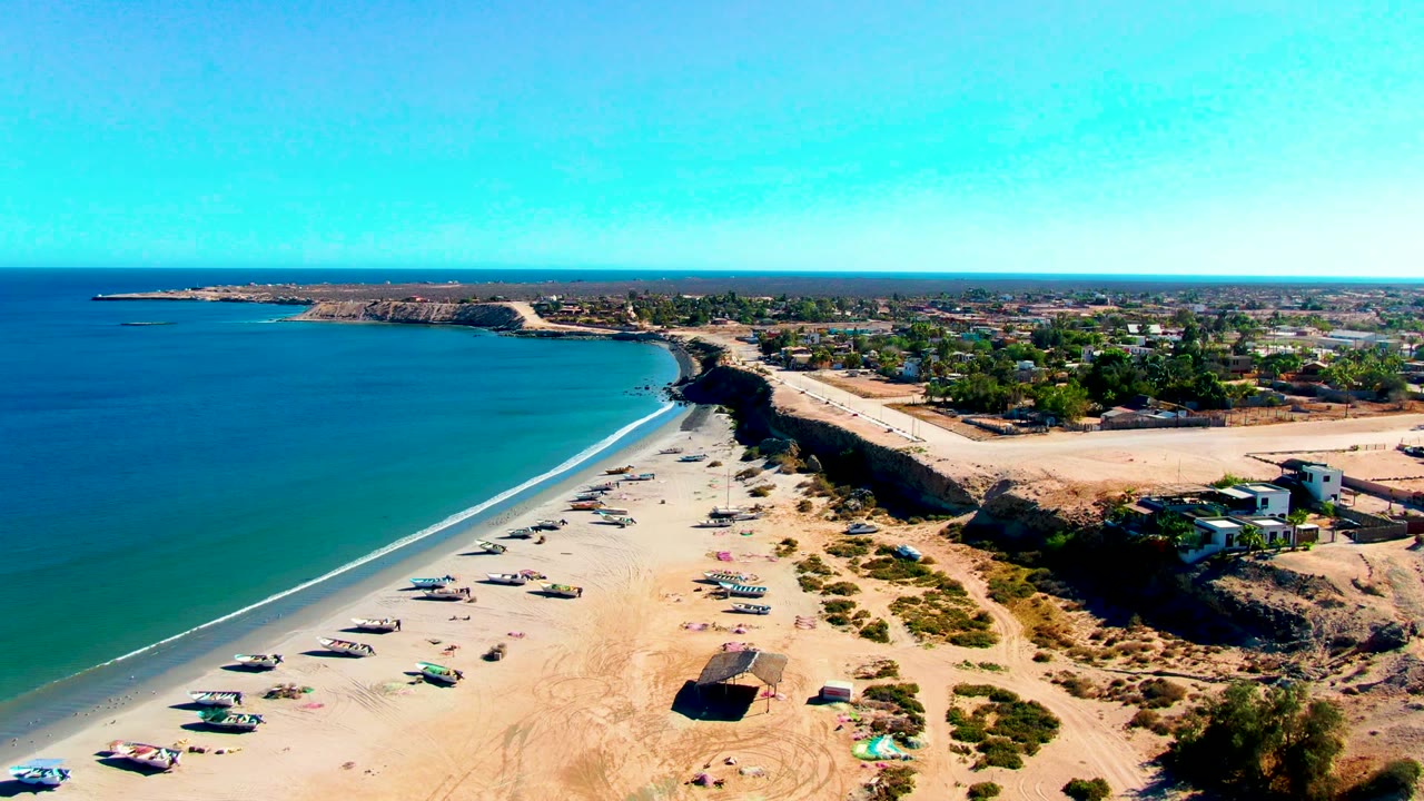 San Juanico aerial view with clear blue ocean, scattered boats pulled onto the sandy beach, low bluffs beside the shore, inland roads and buildings, and no visible people.
