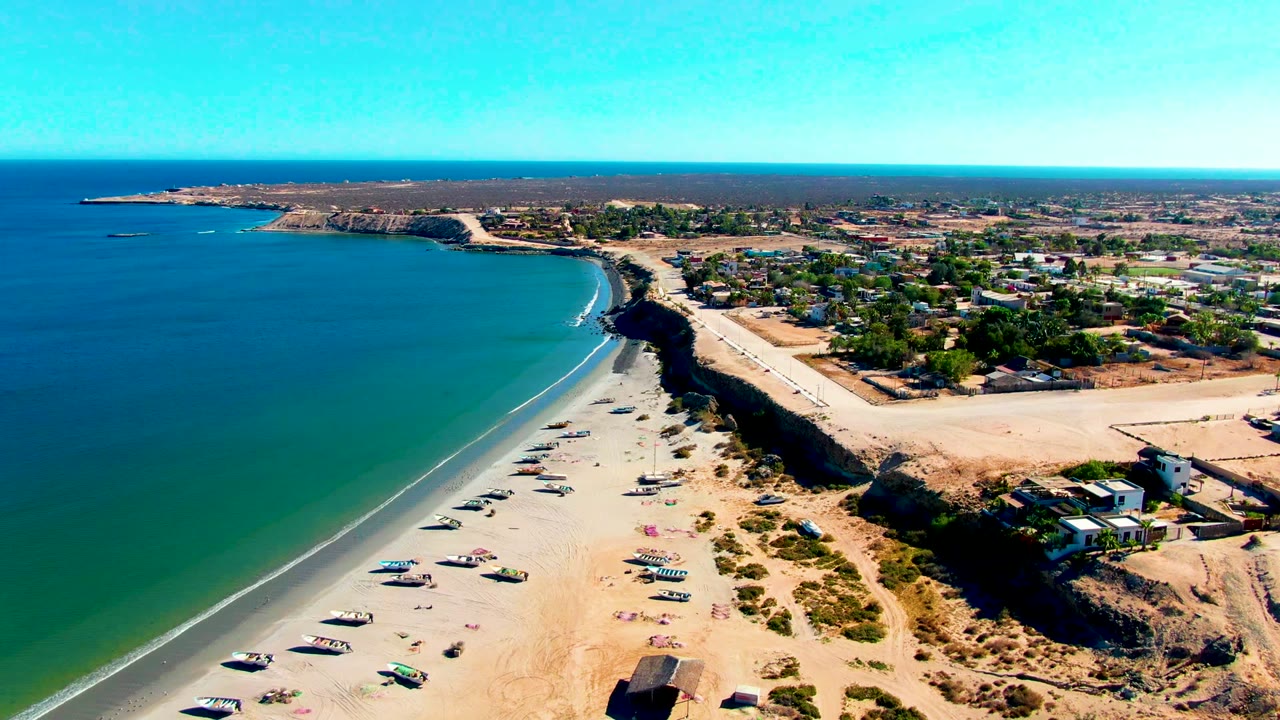 Aerial view over San Juanico, Baja California Sur, showing a curved sandy beach with anchored boats, blue-green water on the left, coastal homes on the right, and no visible people.