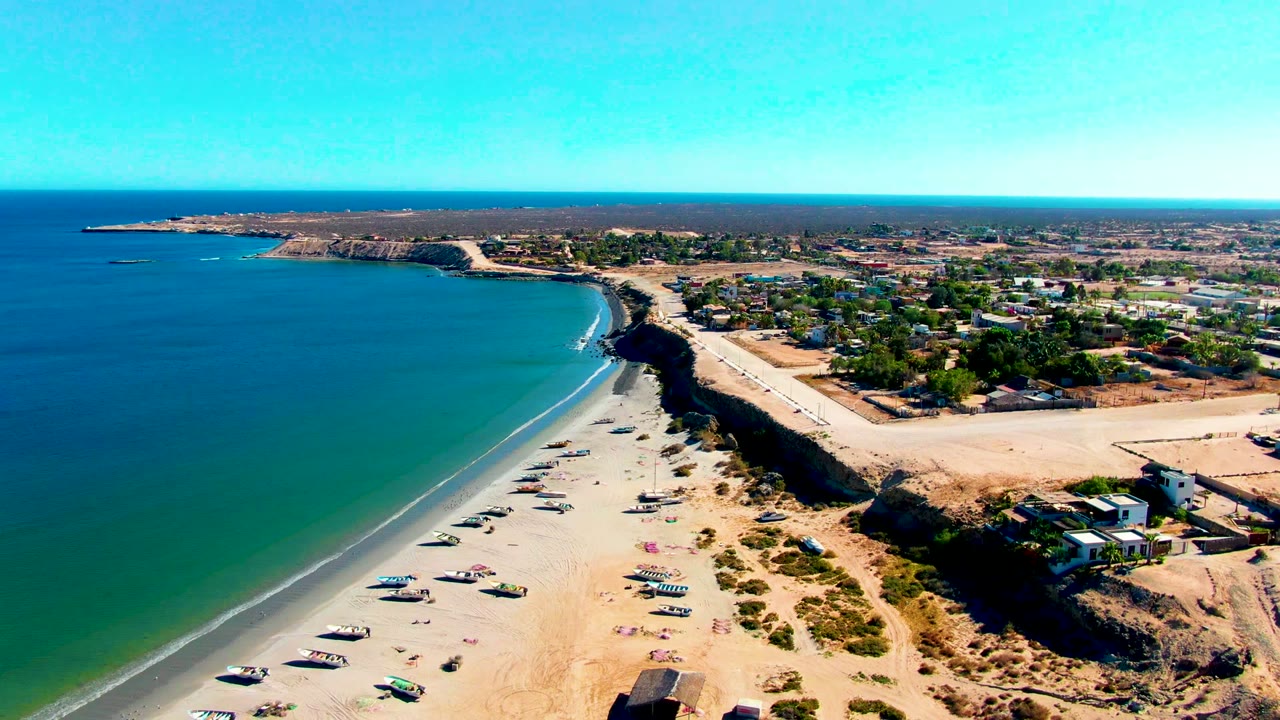 Curving beach with small boats near shore and a white wave line along clear water.