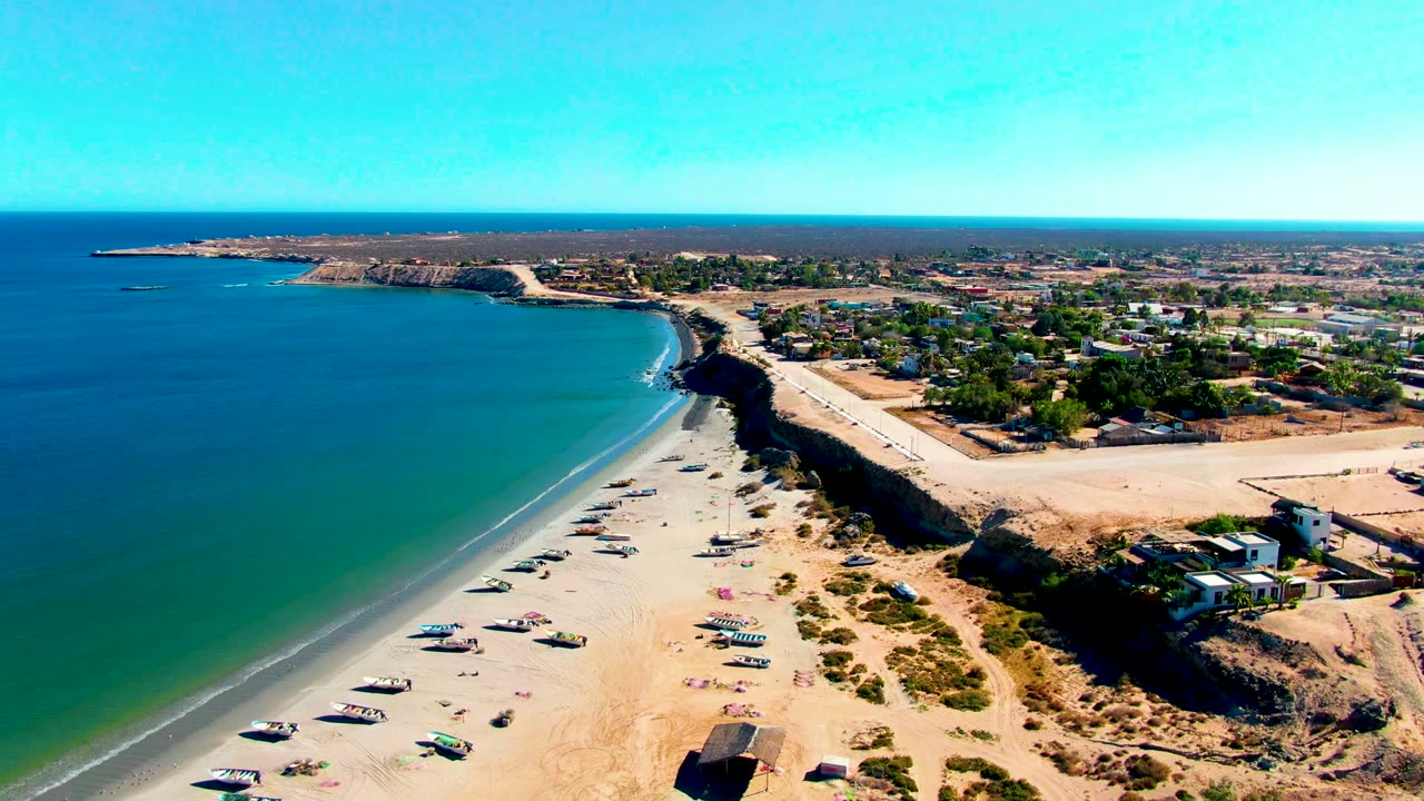 Wide aerial view of San Juanico beach with boats lined across the sand, turquoise water filling the left side, a dark bluff edge, inland houses, and no visible people.