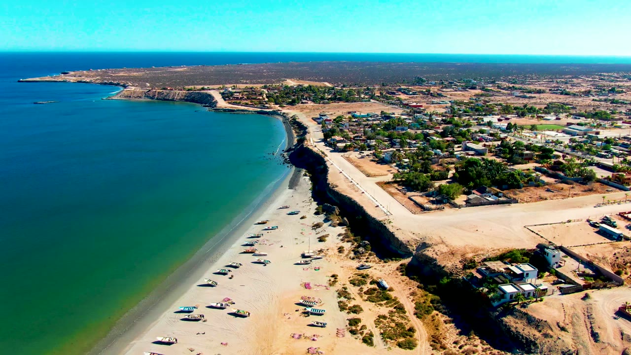 Aerial frame over San Juanico, Baja California Sur showing turquoise ocean, a pale sandy beach with parked vehicles, a dark bluff, nearby buildings, and no visible people.