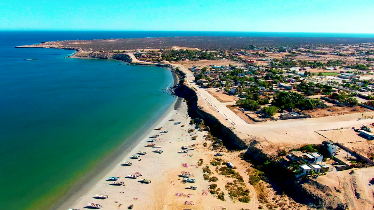 Drone view of sandy shoreline with parked vehicles and low buildings.