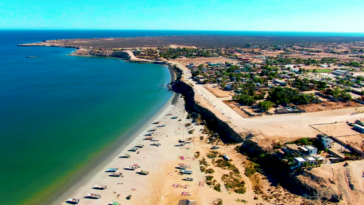 Aerial view over San Juanico with the ocean occupying the left half, beach vehicles near the lower sand, a dark erosion edge, coastal road, inland buildings, and no visible people.