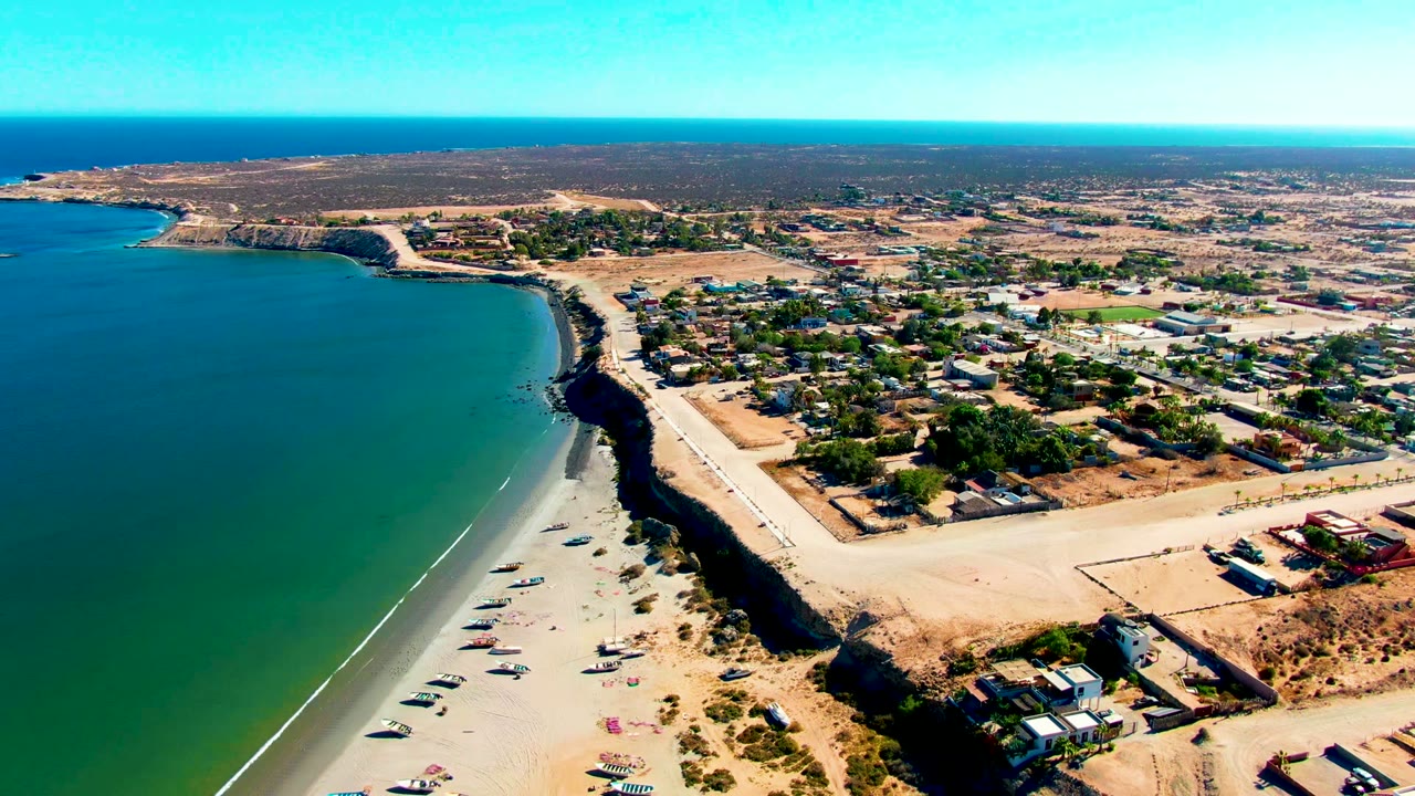 Aerial frame over San Juanico, Baja California Sur, showing green-blue ocean at left, a sandy beach below a dark cliff, a coastal road, town buildings inland, and no visible people.