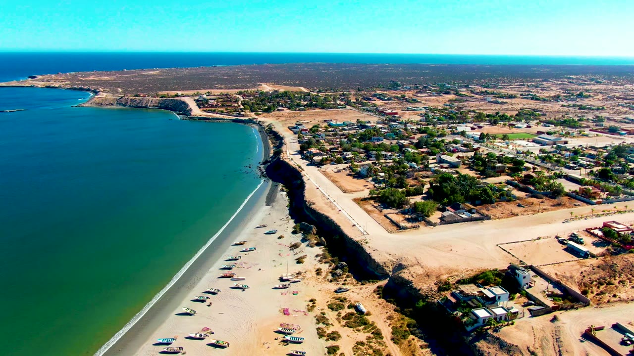 High drone frame over San Juanico, Baja California Sur, showing open ocean to the left, pale sand along the lower edge, a dark bluff beside the road, inland streets and buildings, and no visible people.
