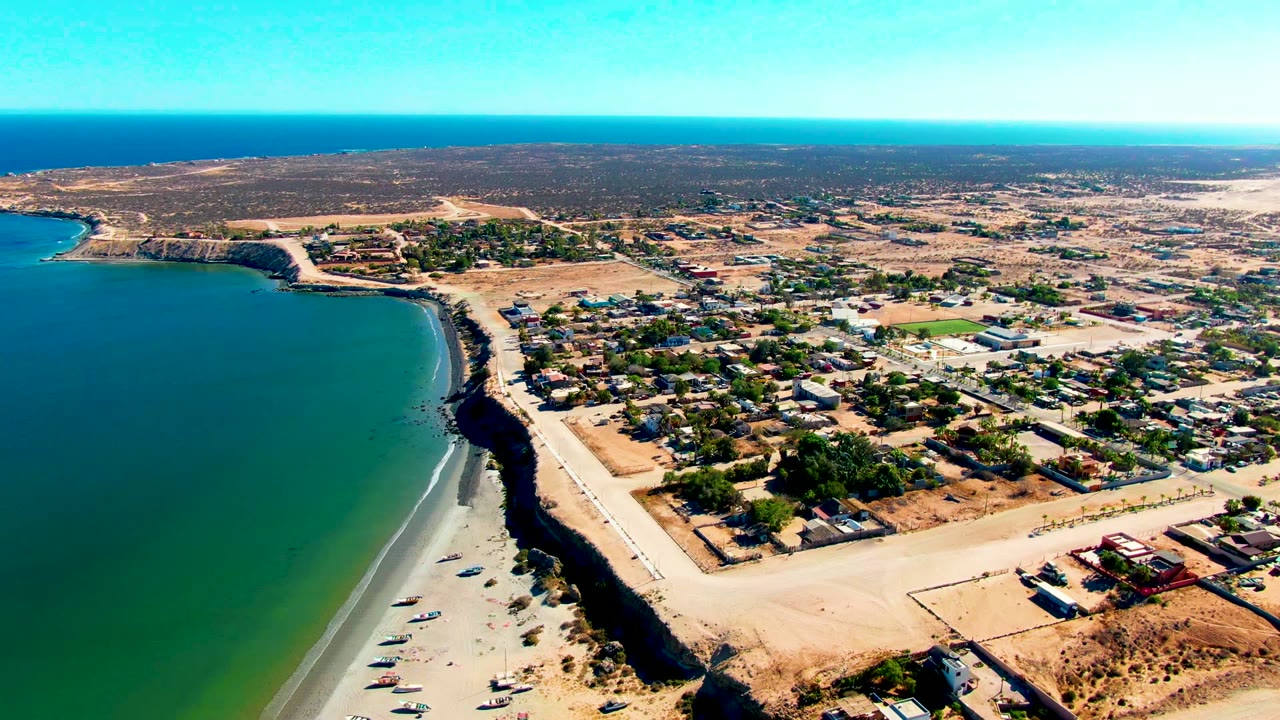 Aerial frame over San Juanico, Baja California Sur, with a curved sandy beach, small boats near shore, town roads and buildings inland, dry scrub beyond, and no visible people.