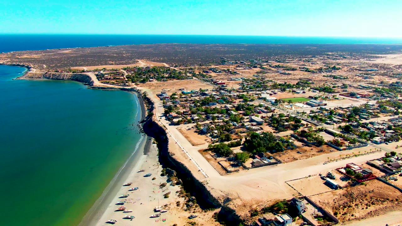 Elevated coastal view with blue water, beach boats, roads, and low buildings.