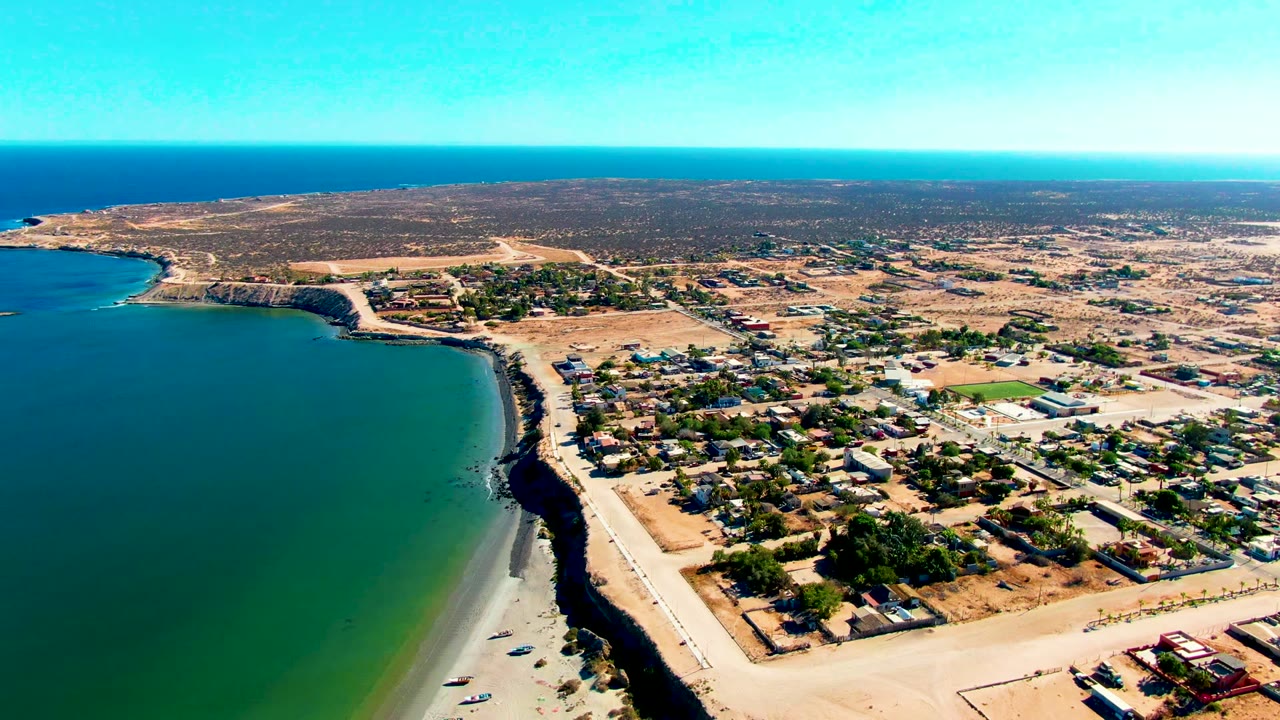 Opening aerial frame over San Juanico, Baja California Sur, with turquoise water at left, a sandy beach below, low buildings inland, dry terrain beyond, and no visible people.