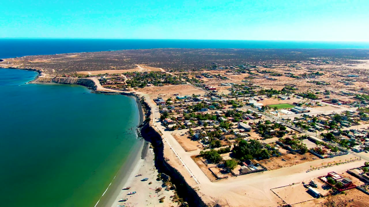 Aerial view of San Juanico with the shoreline and dark coastal edge on the left, beach access roads below, denser building clusters to the right, dry inland terrain, and no visible people.