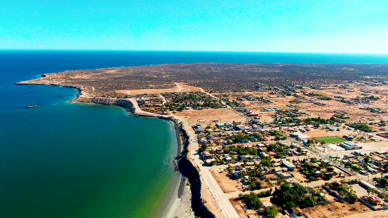 Wide aerial frame over San Juanico, Baja California Sur, with blue-green ocean at left, a sandy beach and road below, low buildings inland, dry terrain beyond, and no visible people.