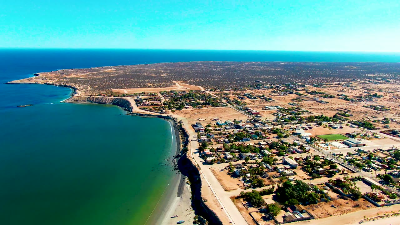 High oblique frame over San Juanico showing ocean on the left, a narrow sandy strip and road along the coast, clustered low structures inland, dry open land behind them, and no visible people.