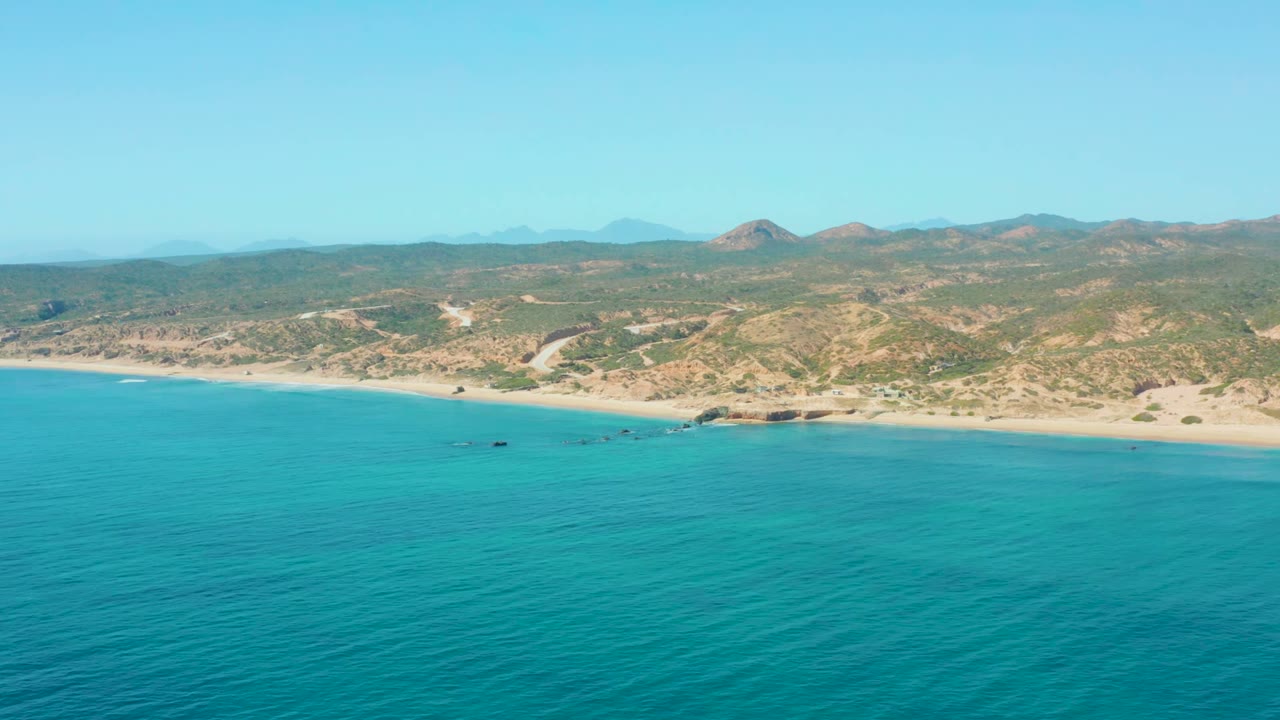 Opening aerial view of El Tule Los Cabos with turquoise water in the foreground, a pale sandy shoreline, scrubby hills, distant mountains, and no visible people.