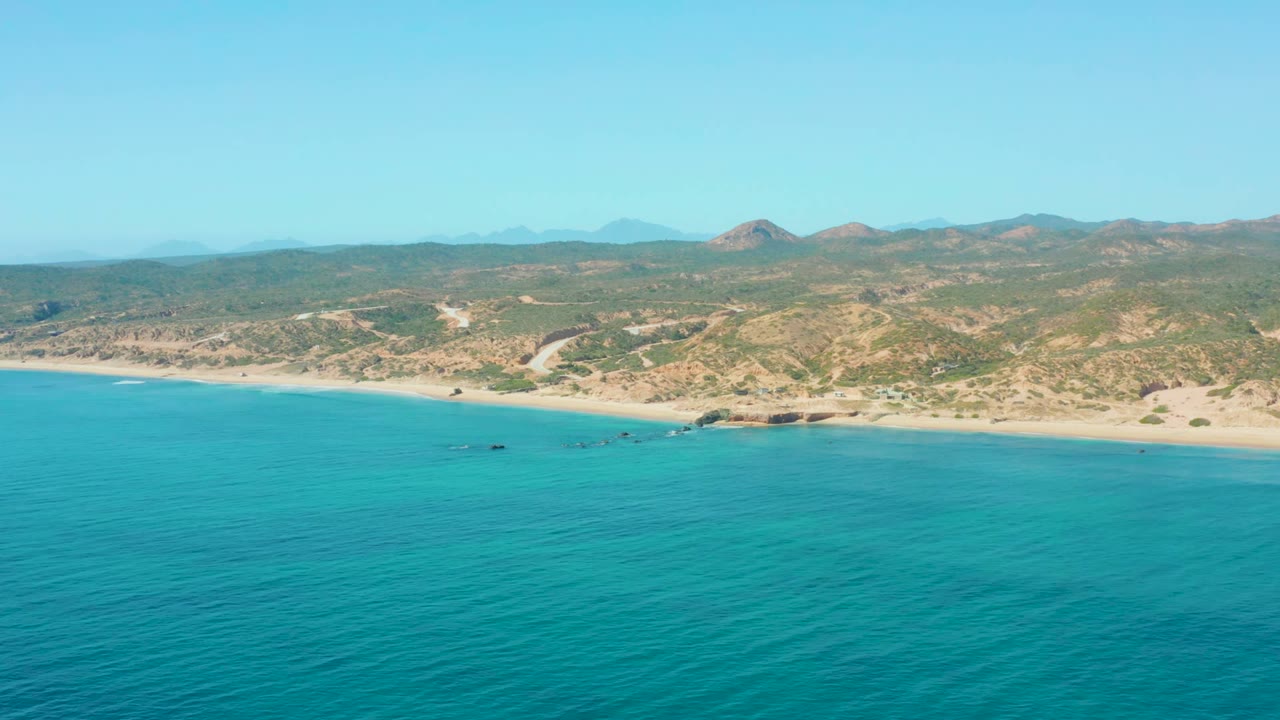 Turquoise coastal water, sandy shoreline, and dry hills under clear sky.
