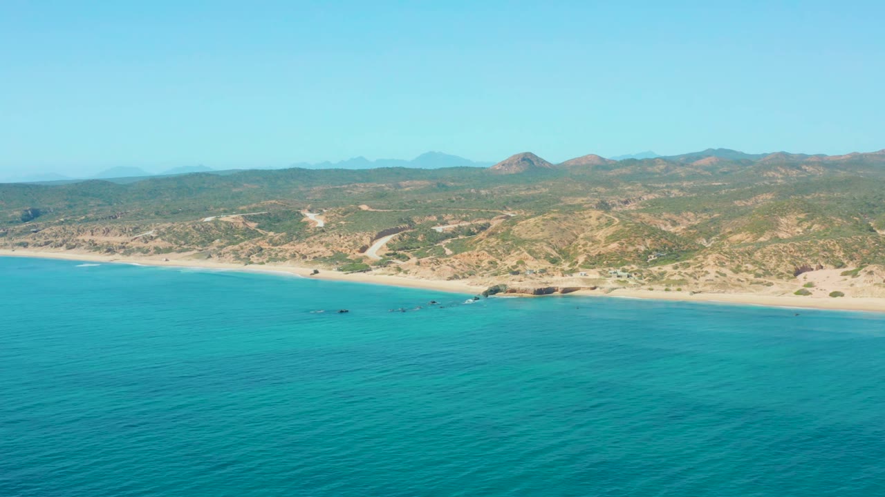 El Tule Los Cabos coastline from above, with textured blue-green ocean across the lower frame, sand and rocks at shore, hillside terrain, and no visible people.