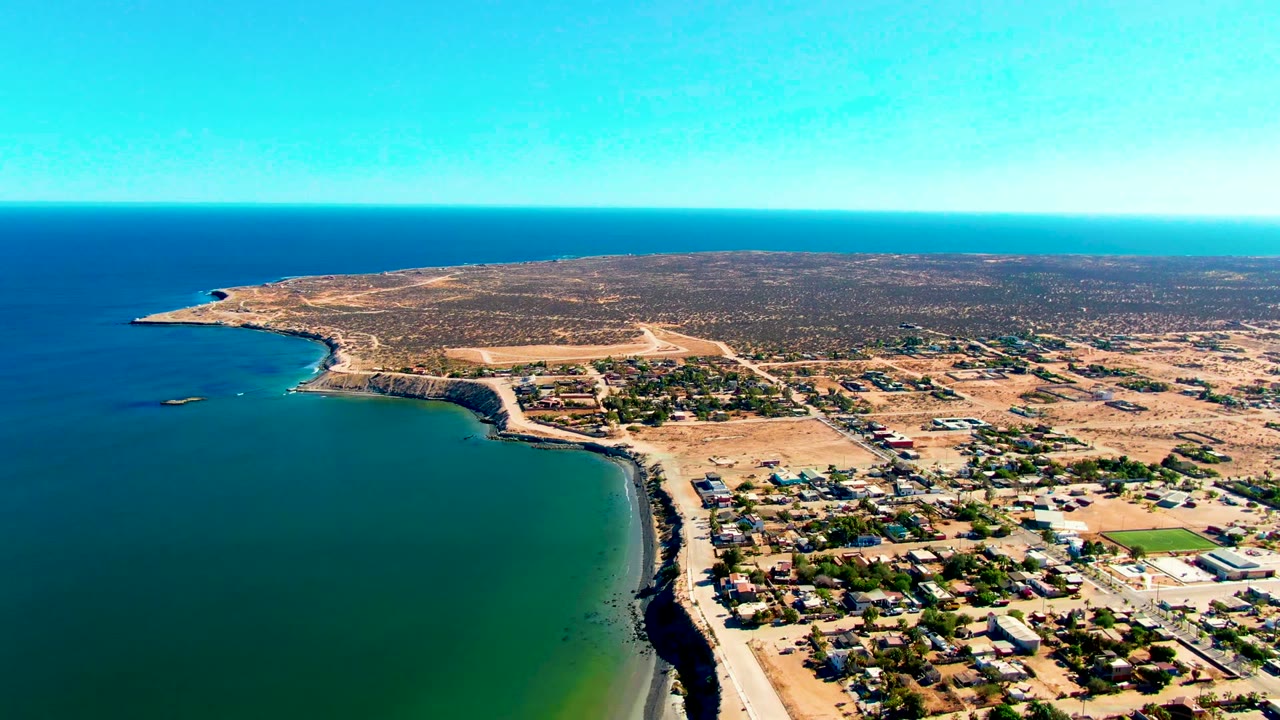 Opening aerial frame over San Juanico, Baja California Sur, with deep blue ocean, a curved rocky shore, dry inland terrain, scattered houses, and no visible people.